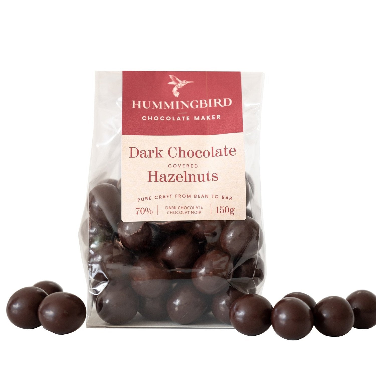 Whole roasted hazelnuts covered in dark chocolate beside wrapped package of Dark Chocolate Covered Hazelnuts by Hummingbird Chocolate Maker