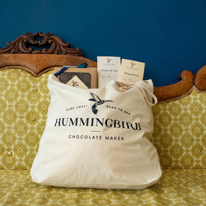 Hummingbird Chocolate Tote Bag resting on ornate yellow couch filled with chocolate bars and caramels